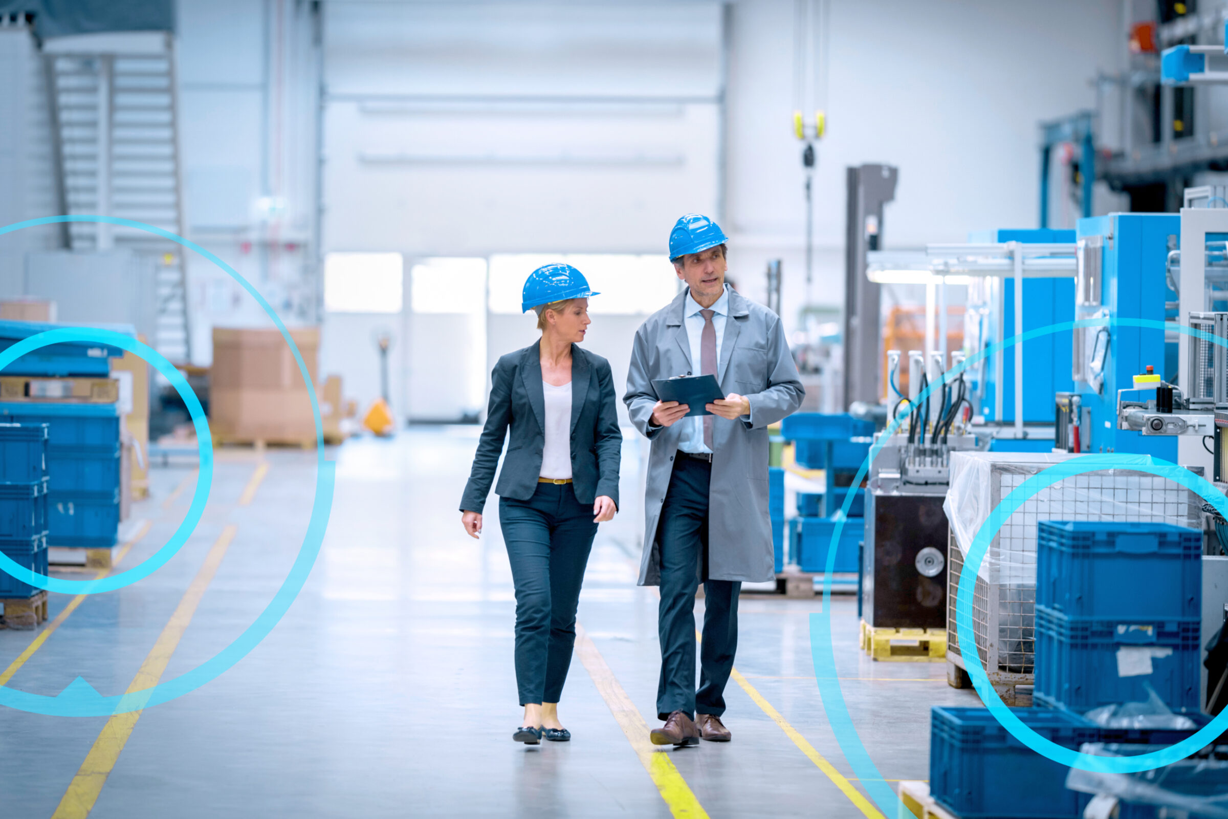 There are two people walking through a factory with a tablet an helmets on.